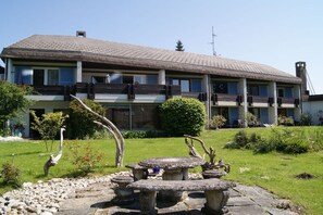 Un bâtiment à deux étages, doté d’un toit plat, d’un balcon à l’étage supérieur et d’une table en pierre avec des chaises dans le jardin avant.