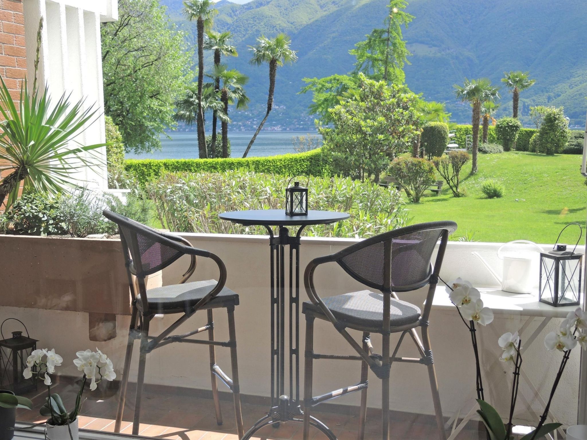 Een balkon met twee rieten stoelen en een kleine tafel, met uitzicht op een verzorgde tuin en een waterpartij met bergen op de achtergrond.