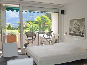 A bedroom with a bed, two chairs, a table, and a view of a garden and mountains. - San Remo by Interhome (Brissago)