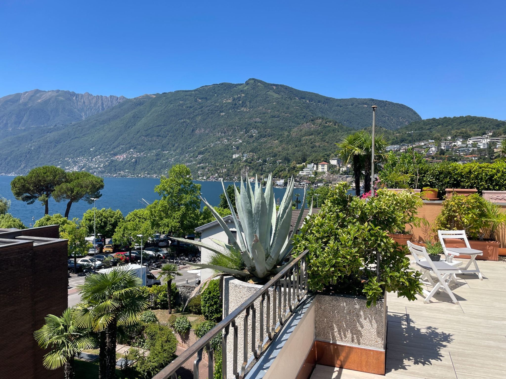 A rooftop terrace with a view of the sea and mountains, featuring a large agave plant, outdoor seating, and a parking area.