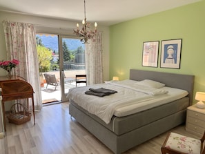 Una camera da letto con un letto, un comodino, una sedia, un tavolo con un vaso di fiori e vista sull'esterno.