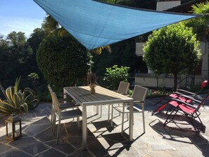 Un patio al aire libre con una mesa y sillas, una vela de sombra azul y diversas plantas.