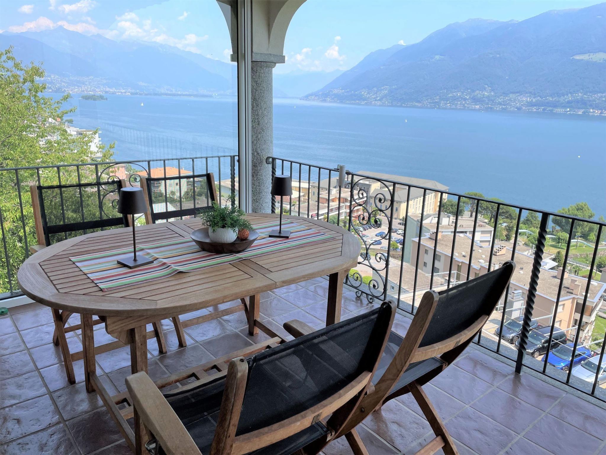 Une table en bois, des chaises et une plante en pot sur un balcon offrant une vue sur un lac et des montagnes.