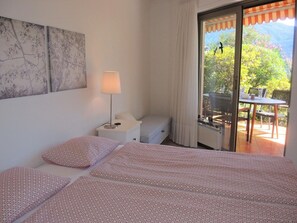 A bedroom with a bed, a nightstand, a lamp, and a view of an outdoor patio with a table and chairs. - Aphrodite by Interhome (Brissago)