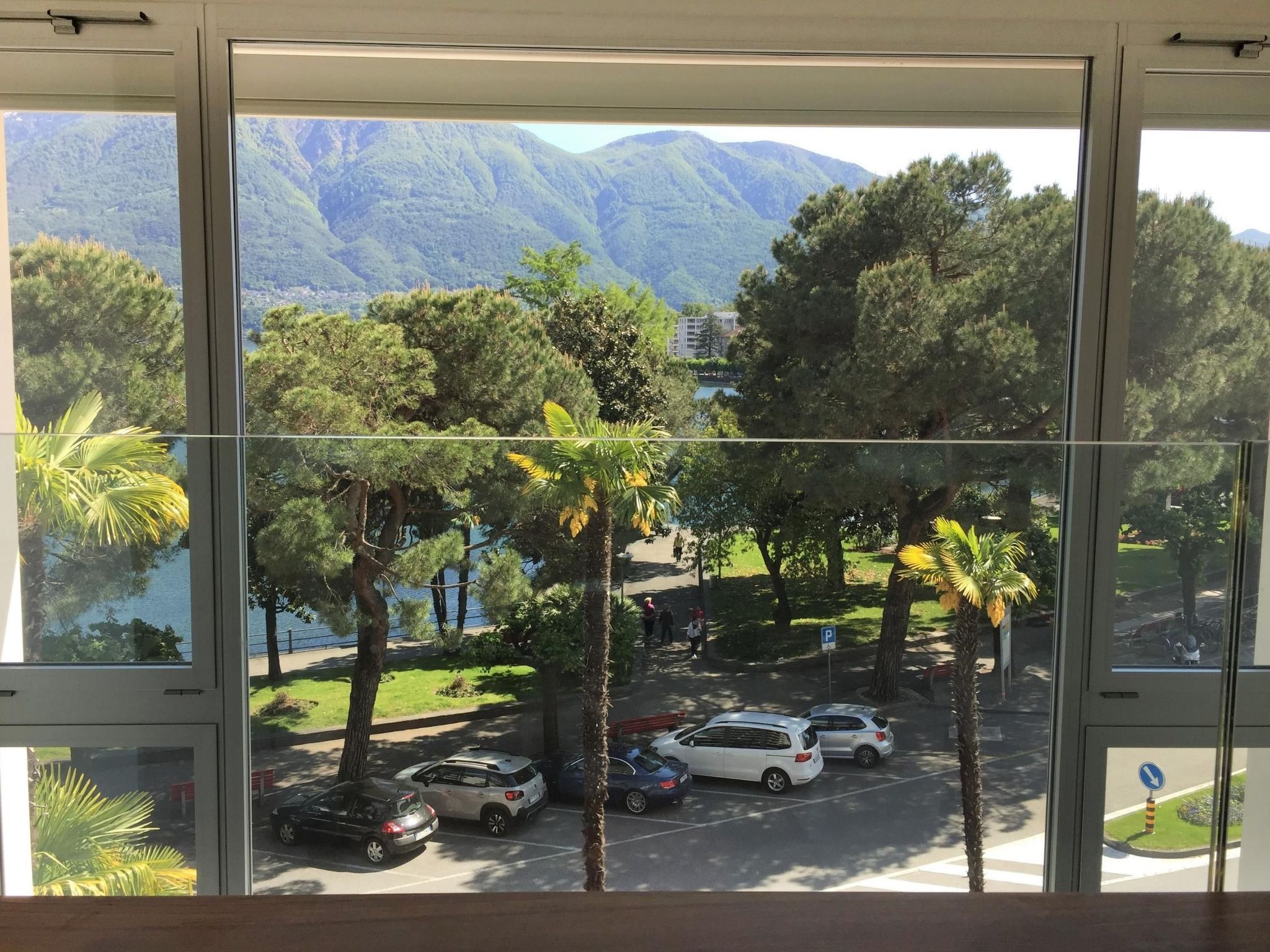 Une vue à travers une fenêtre sur un parking où l’on aperçoit des voitures, des palmiers et, au loin, un paysage montagneux.