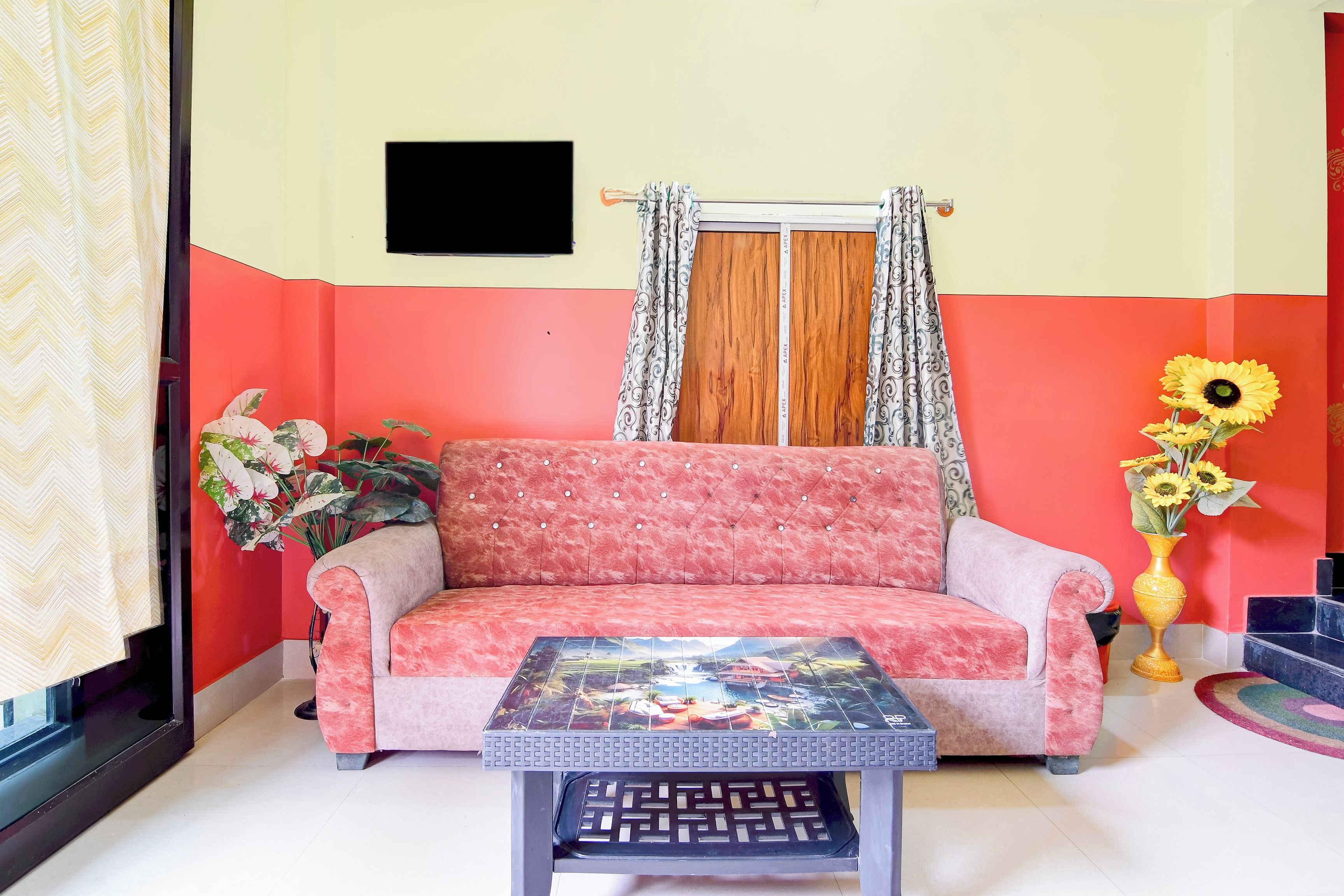 A room with a sofa, a table, a window with curtains, a wall-mounted TV, and a vase with flowers.
