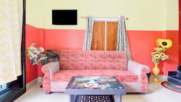 Una habitación con sofá, una mesa, un ventanal con cortinas, un televisor montado en la pared y un jarrón con flores.