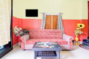 A room with a sofa, a table, a window with curtains, a wall-mounted TV, and a vase with flowers.