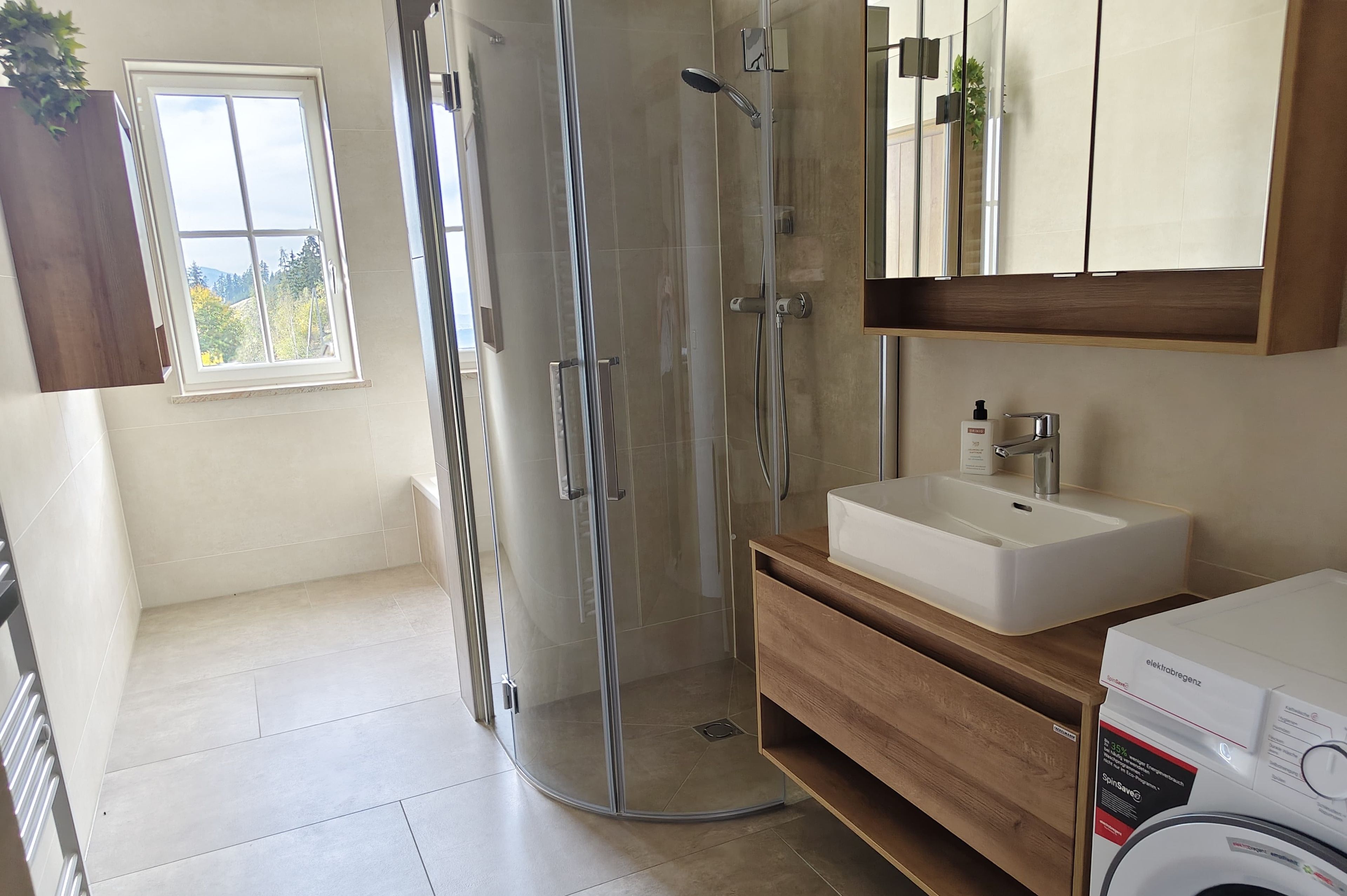 Une salle de bain moderne avec une douche à l’italienne en verre, un lavabo avec un meuble-vasque en bois et une fenêtre donnant sur les arbres.