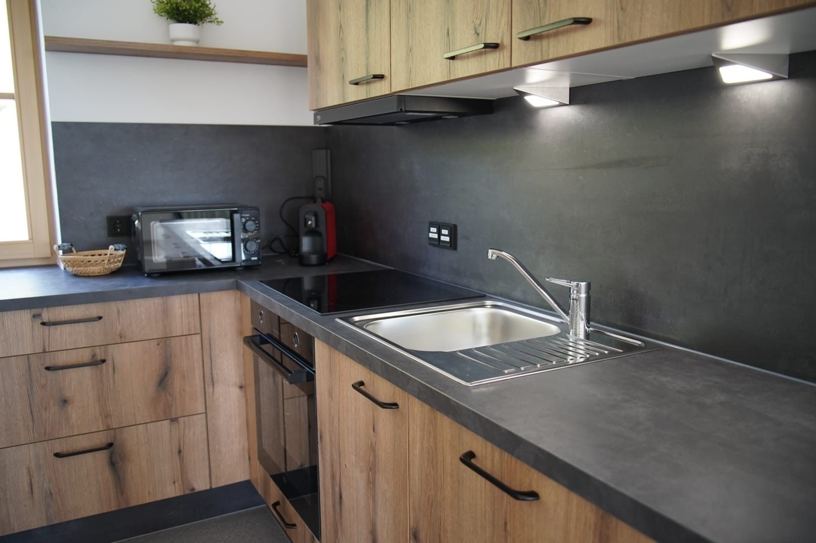 Une cuisine avec des meubles de rangement en bois, un plan de travail noir, un évier en acier inoxydable, un four à micro-ondes et une bouilloire.