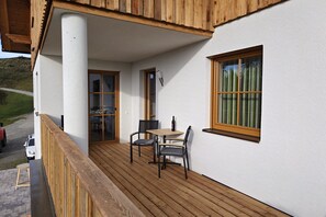 Een houten terras met een tafel en stoelen, een raam met gordijnen, en een deur naar de binnenruimte.