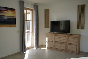 Ein Zimmer mit großem Fenster, einem Flachbildfernseher auf einer Holzkommode und einem Gemälde an der Wand.