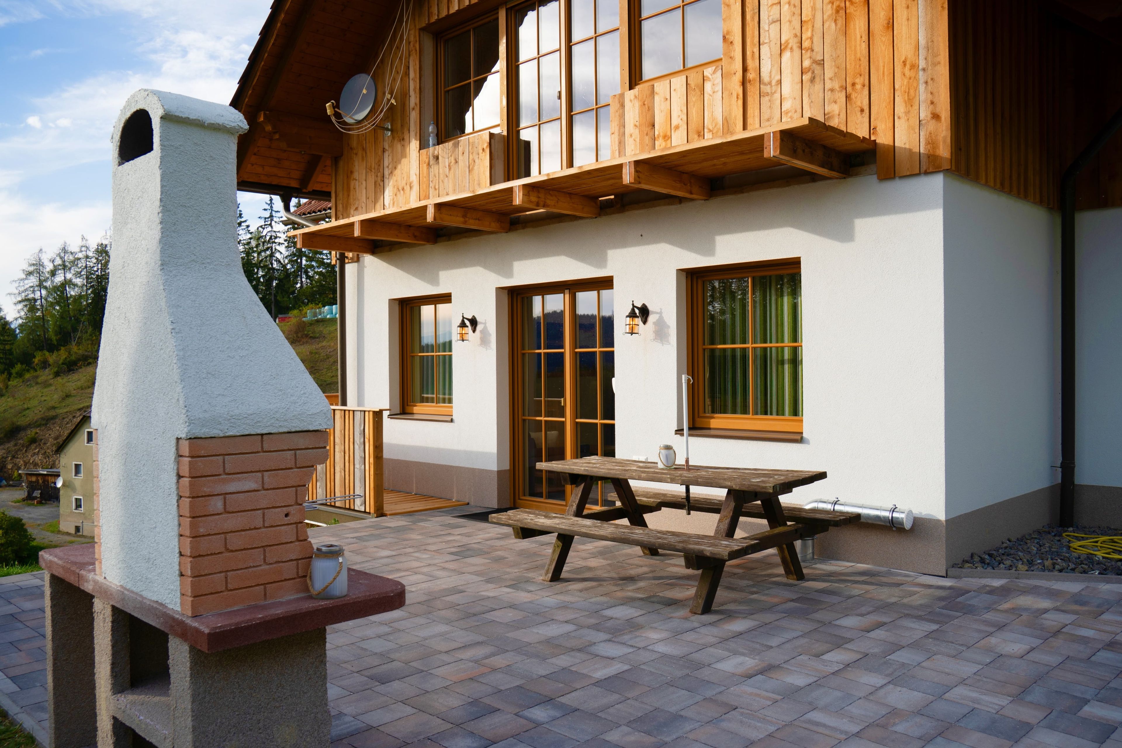 Une maison en bois avec une cheminée, une terrasse avec une table et des bancs, et une fenêtre donnant sur le paysage environnant.