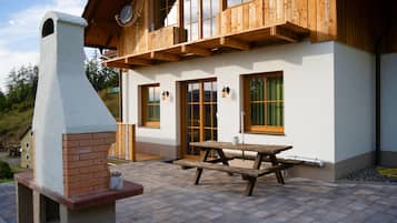 Une maison en bois avec une cheminée, une terrasse avec une table et des bancs, et une fenêtre donnant sur le paysage environnant.