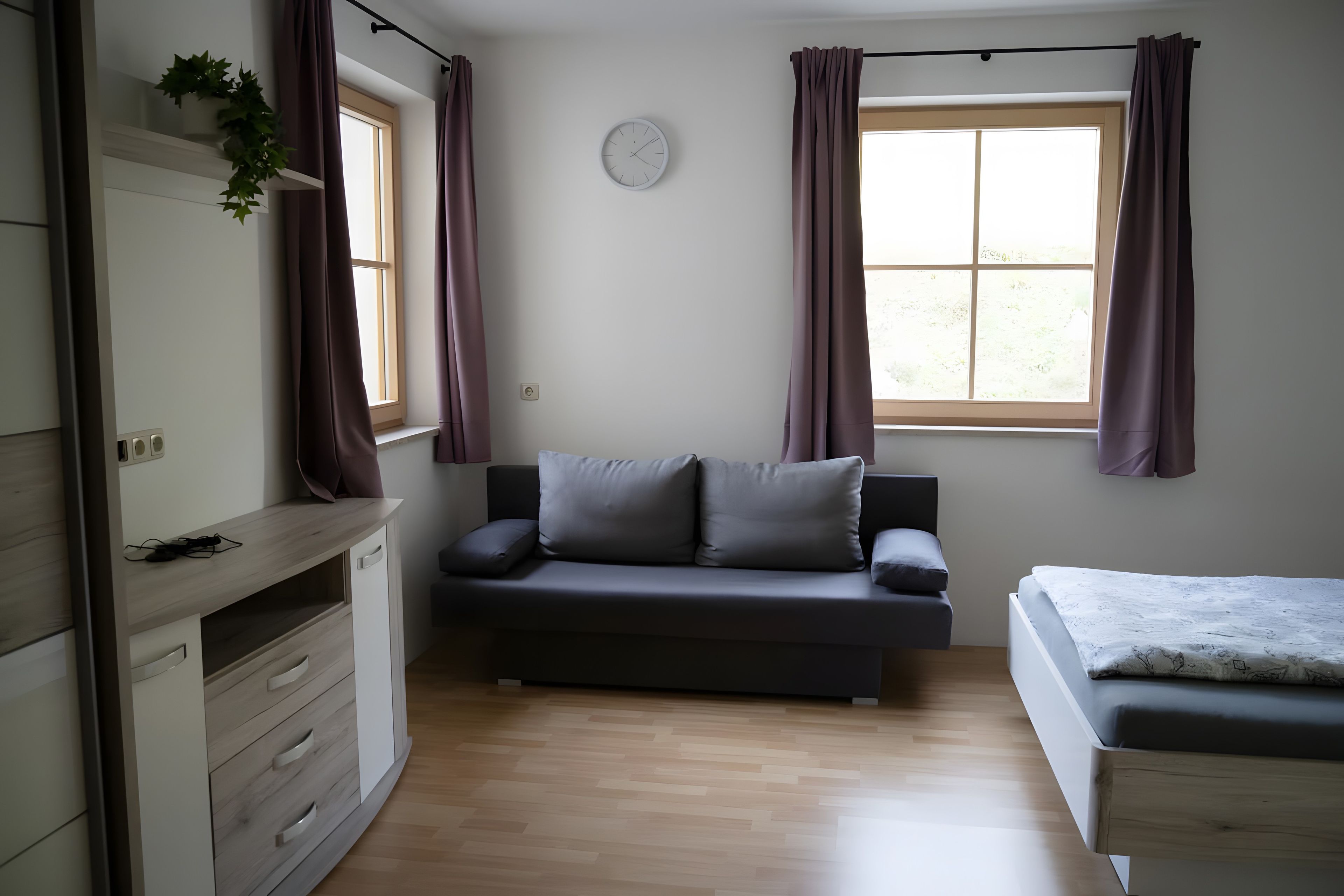 Et moderne, minimalistisk værelse med en sofa, en seng, et skrivebord og et vindue med gardiner.