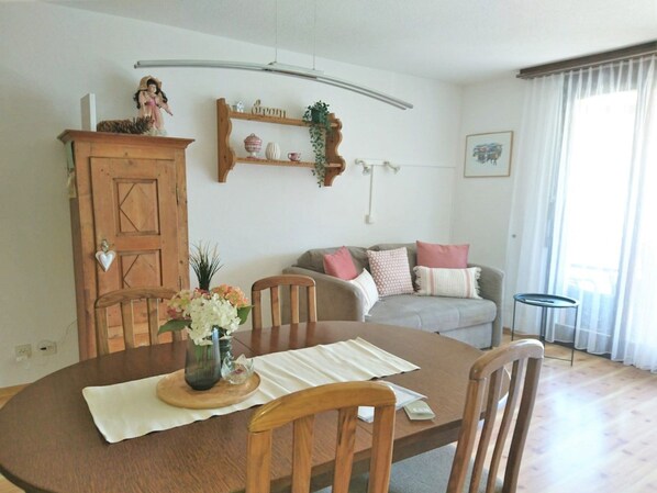En matplats med ett träbord, stolar och en vas med blommor. I bakgrunden finns en soffa och ett skåp.