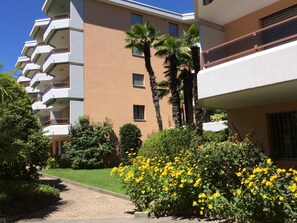 Un immeuble à plusieurs étages doté de balcons, entouré d’un espace verdoyant bien entretenu et de plantes à fleurs jaunes.
