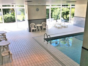 Un espace piscine intérieure doté d’un sol carrelé, d’une piscine, ainsi que de chaises et de tables en plastique blanc.