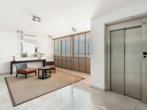 A modern elevator with a metallic finish, a small seating area with two chairs and a table, and a large window with blinds. - Residenza Acapulco I by Interhome (Porto Ronco)