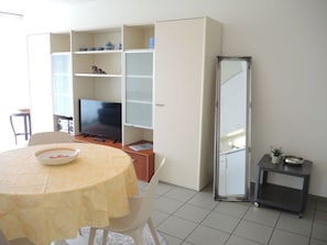 A living room with a TV, a dining table with a bowl, a cabinet with shelves, a mirror, and a small table with a plant. - San Materno by Interhome (Ascona)