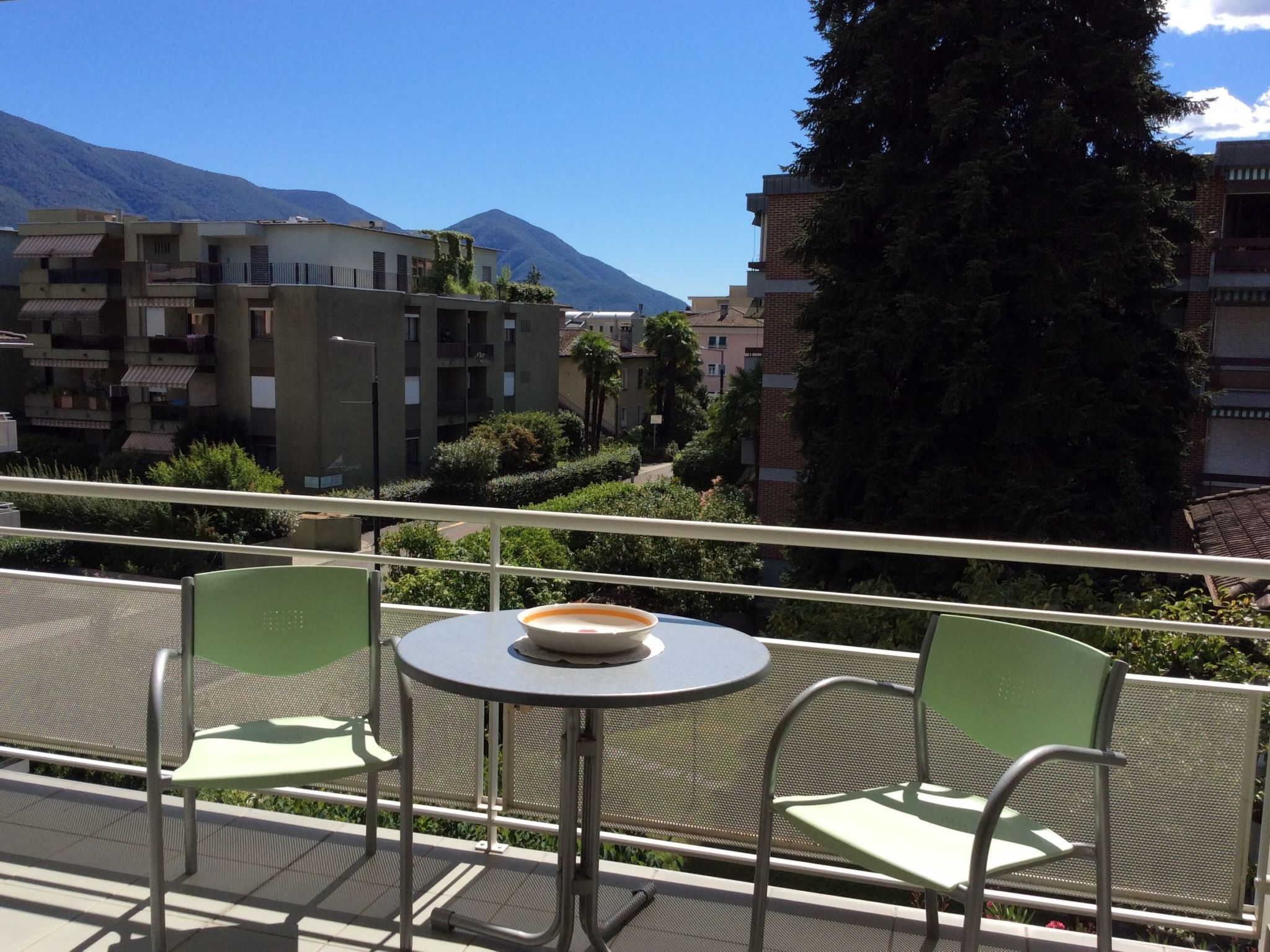 A balcony with a round table and two chairs, overlooking a residential area with buildings and mountains.
