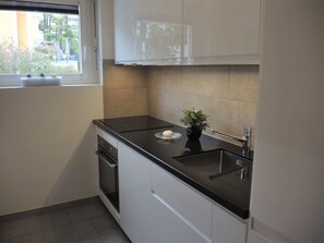A kitchen with white cabinets, a black countertop, a sink, and a window with a view of greenery outside. - Appartement Violetta by Interhome (Ascona)