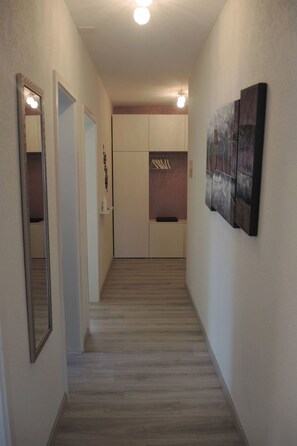 Een gang met een houten vloer, een deur aan de linkerkant en een wand met een ingelijste foto en een lamp.