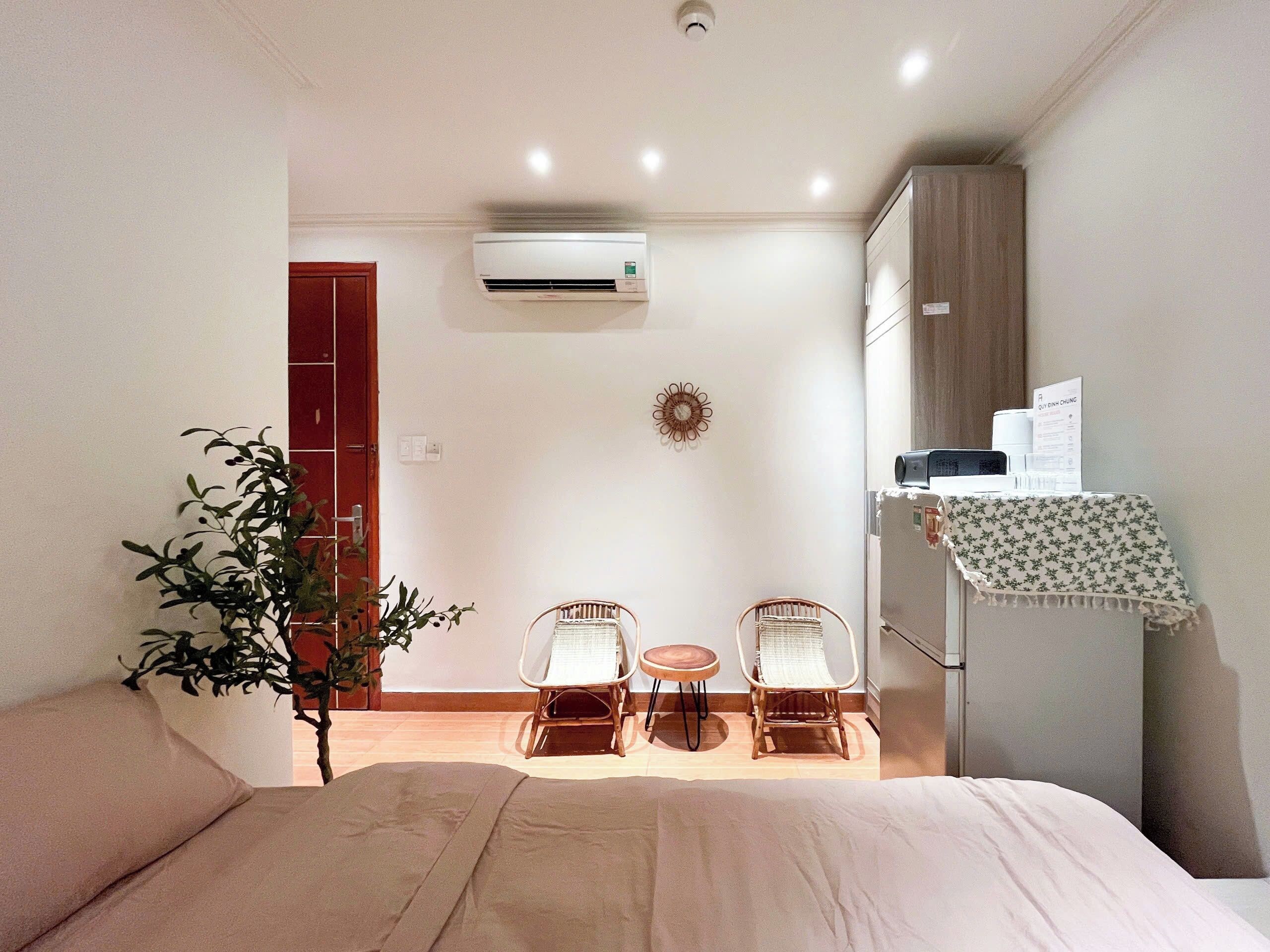 A hotel room with a bed, two chairs, a small table, an air conditioning unit, a wall-mounted clock, and a door.