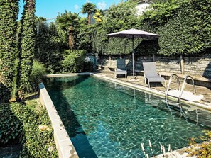 A clear swimming pool with a stone wall, surrounded by lush greenery and a stone bench. - Testa Nera by Interhome (Brione sopra Minusio)