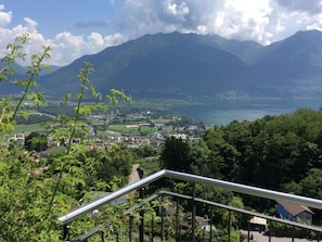 A panoramic view of a mountainous landscape with a lake, a town in the valley, and a metal railing in the foreground. - Casa Lucciola by Interhome (Tenero-Contra)