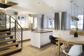 A modern kitchen with a central island, a dining area with a table and chairs, and a staircase with a stone accent wall. - Heimat1883 - Schofpauli (Garmisch-Partenkirchen)