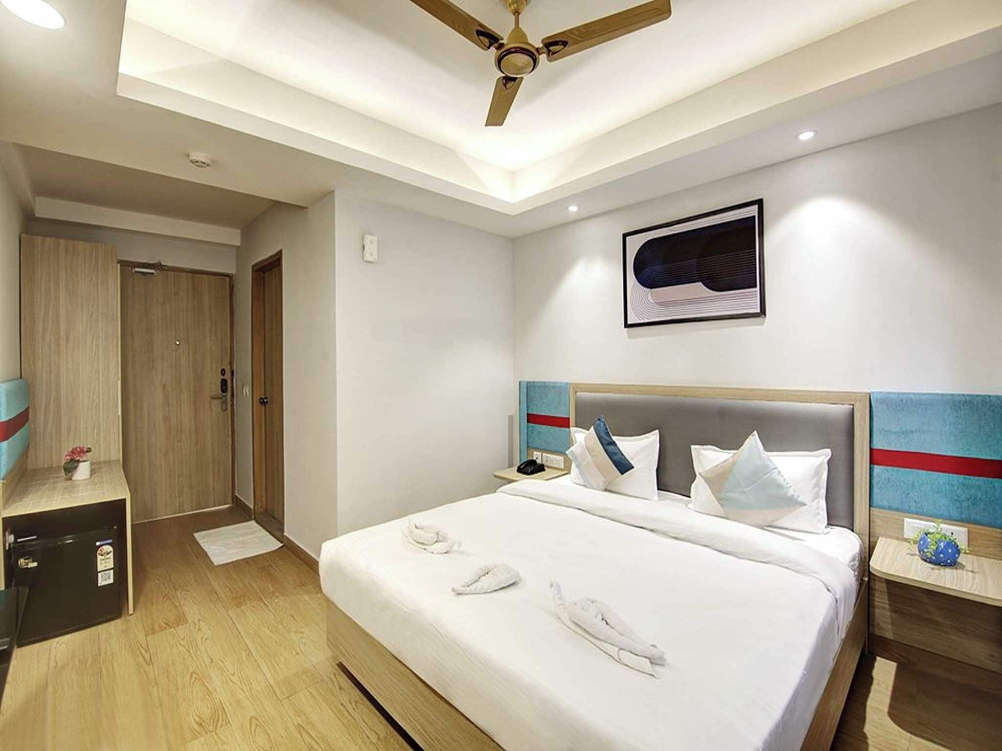 A hotel room with a bed, a ceiling fan, a framed artwork, and a wooden wardrobe.
