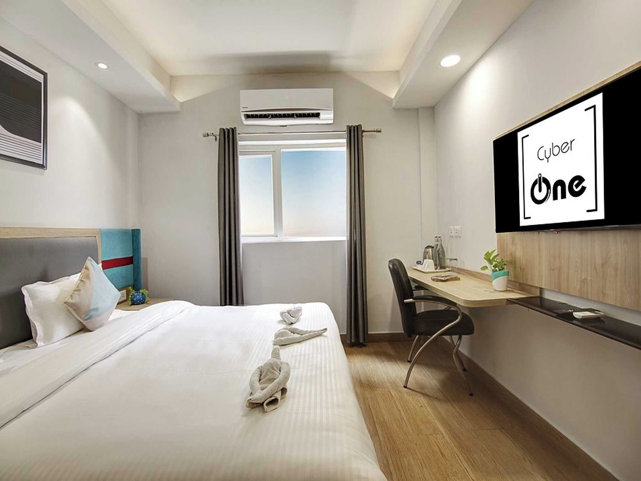 A hotel room with a bed, a desk, a chair, an air conditioning unit, a window with curtains, and a TV screen displaying 'Cyber One'.
