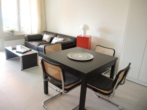 Ein modernes Wohnzimmer mit einer schwarzen Ledersofa, einem roten Beistelltisch, einem Esstisch mit vier Stühlen und einer Stehlampe.
