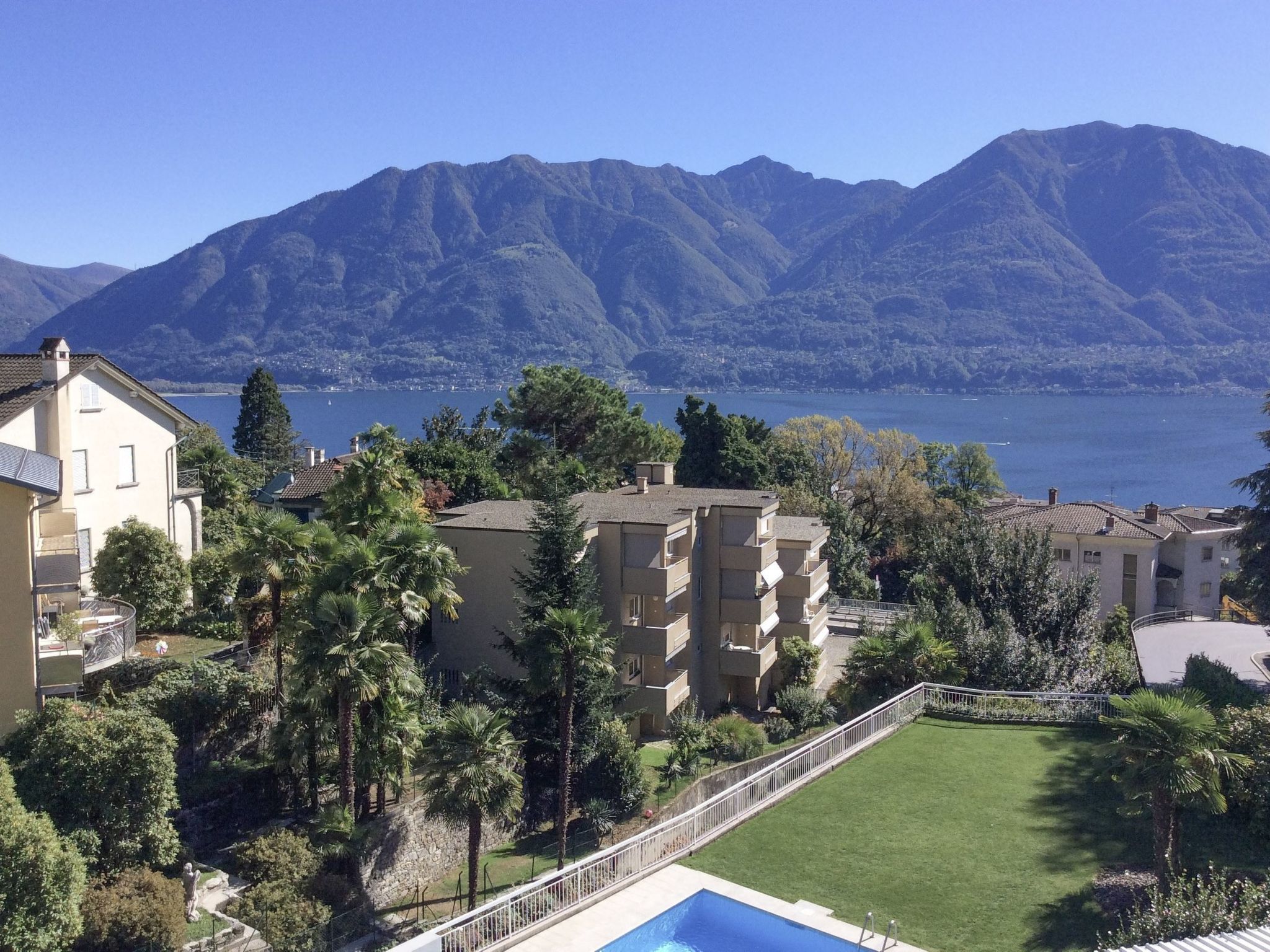 A residential area with a swimming pool, surrounded by greenery and overlooking a lake with mountains in the background.