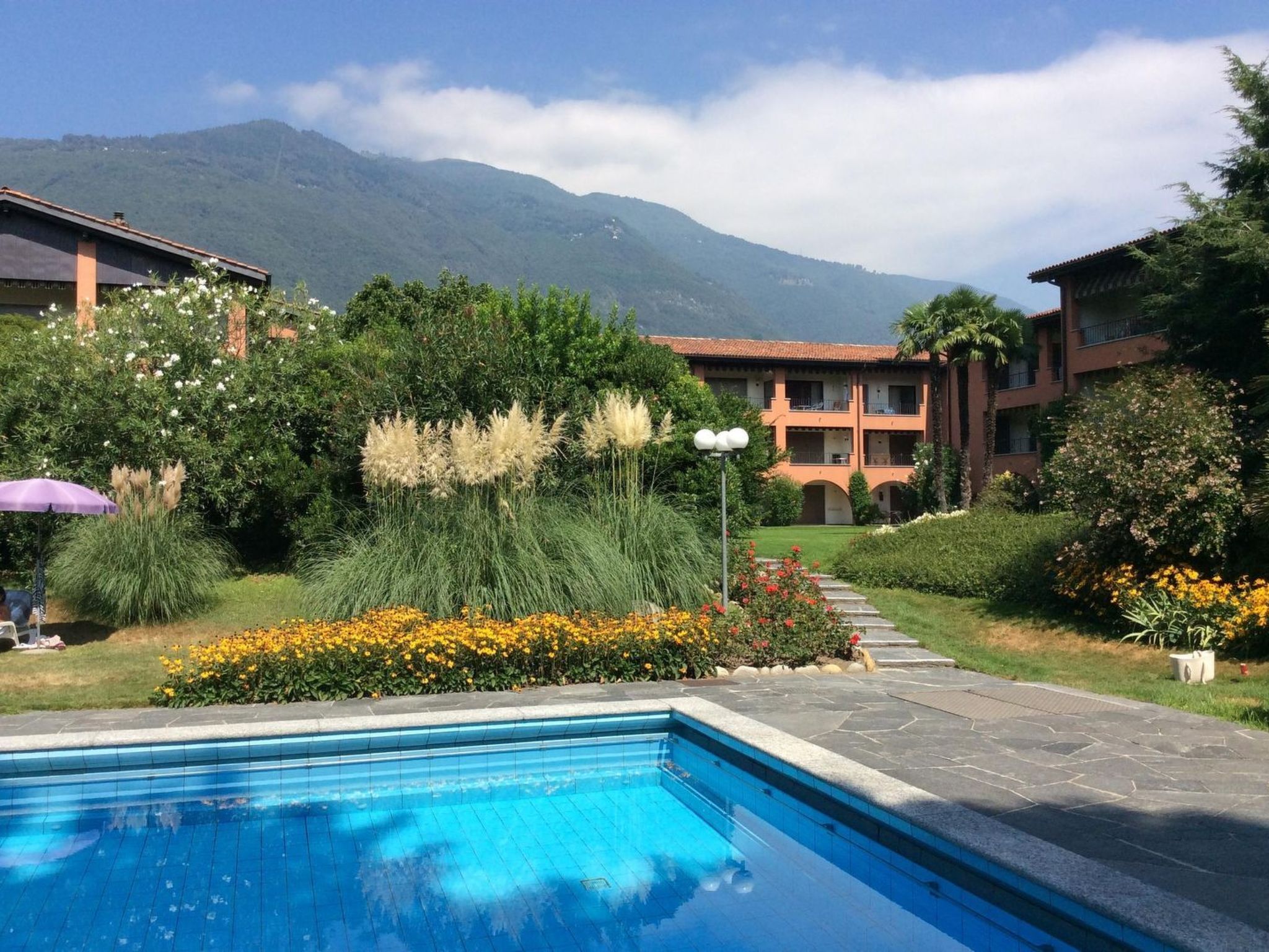 A swimming pool with a mountain backdrop, surrounded by well-maintained gardens and a building with multiple balconies.