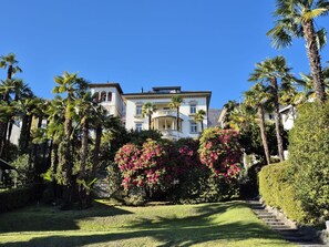 A large, multi-story building with a central courtyard, surrounded by palm trees and flowering bushes. - Villa Clara by Interhome (Minusio)