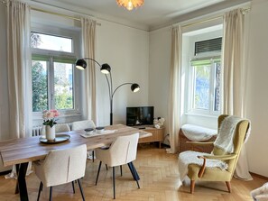 A modern living room with a wooden dining table, chairs, a television, and a large window with curtains. - Villa Clara by Interhome (Minusio)