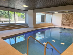 A modern indoor swimming pool with clear glass walls, a wooden ceiling, and a view of greenery outside. - Portofino by Interhome (Brissago)