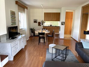 A modern living room with a flat-screen TV, a dining area with a table set for two, and a kitchen with wooden cabinets. - Al Lido by Interhome (Locarno)