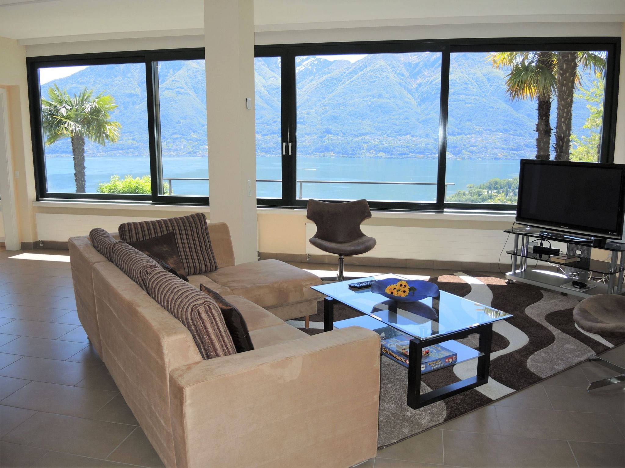 Una sala de estar moderna con un sofá, una mesa de centro y un televisor. Ventanas amplias ofrecen vistas a montañas y un lago.