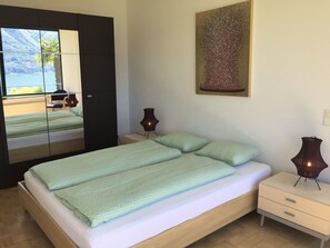 A bedroom with a bed, bedside tables, a mirror, a painting, and a view of the outdoors. - Monte Fiorito by Interhome (Orselina)