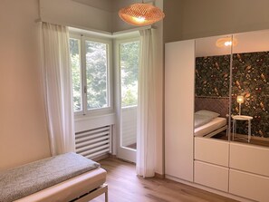 A bedroom with a bed, a window with curtains, a radiator, a wardrobe, and a mirror with a decorative wall. - Casa Colombina Duplex by Interhome (Gordola)