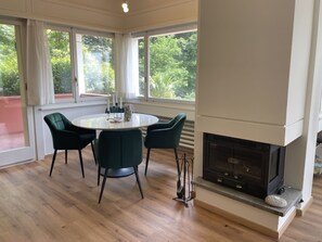 A modern dining area with a round table, four chairs, a fireplace, and large windows with curtains. - Casa Colombina Duplex by Interhome (Gordola)