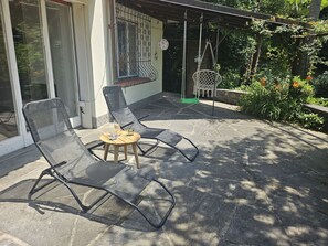 Une terrasse extérieure avec deux chaises longues et une petite table, entourée d’un sol en pierre et de verdure.