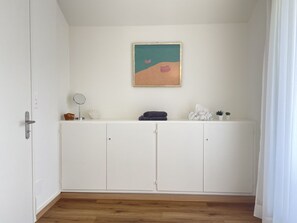 Ein Zimmer mit einem weißen Kleiderschrank, einer kleinen Ablage mit Spiegel und einem Bild an der Wand.