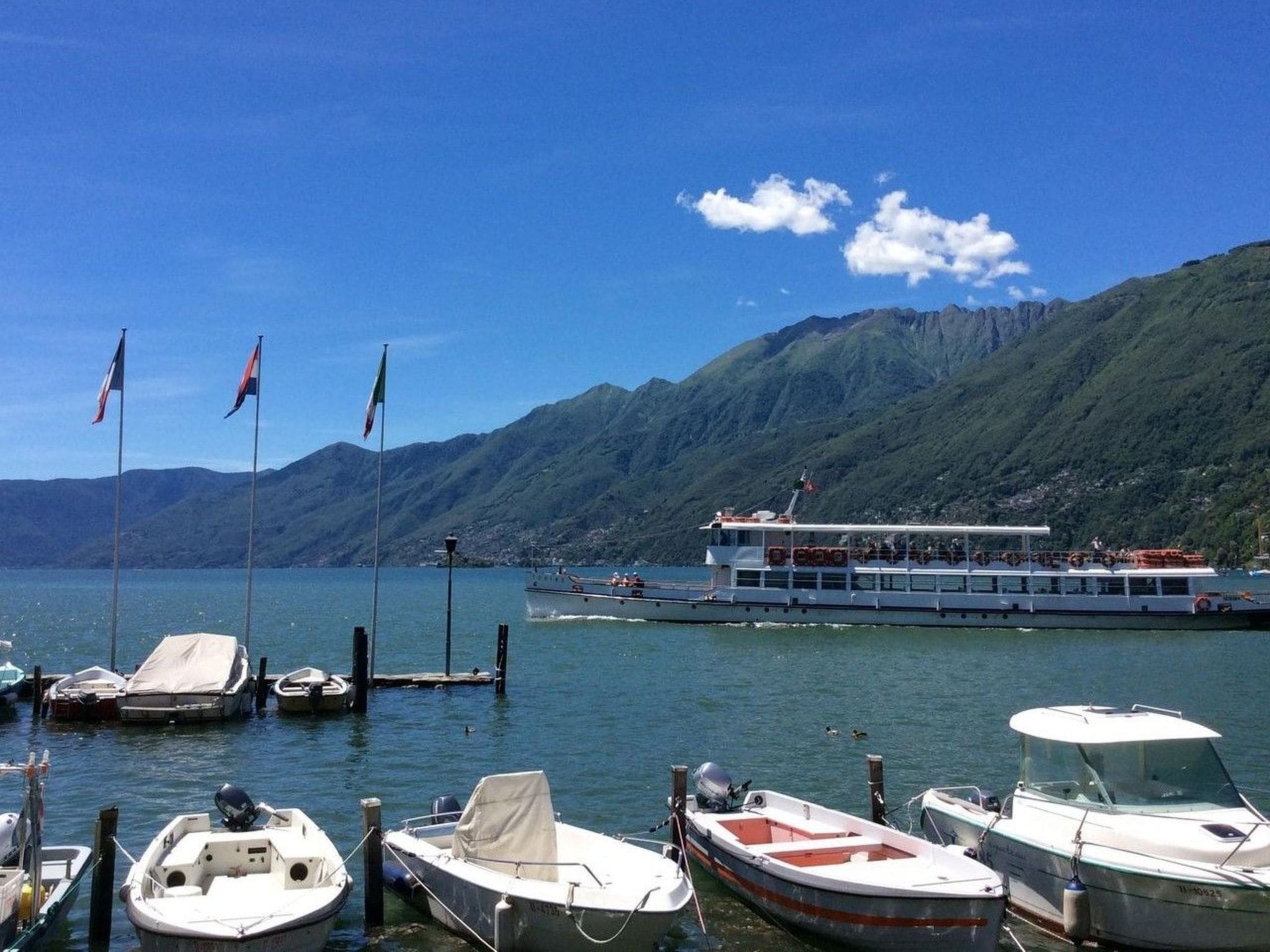 Un port de plaisance où sont amarrés plusieurs bateaux, un grand ferry, et des montagnes en arrière-plan.