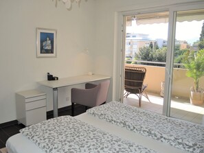 Ein Hotelzimmer mit Bett, Schreibtisch, Stuhl, kleinem Tisch, einem gerahmten Bild an der Wand und einem Balkon mit Blick auf Gebäude und Bäume.