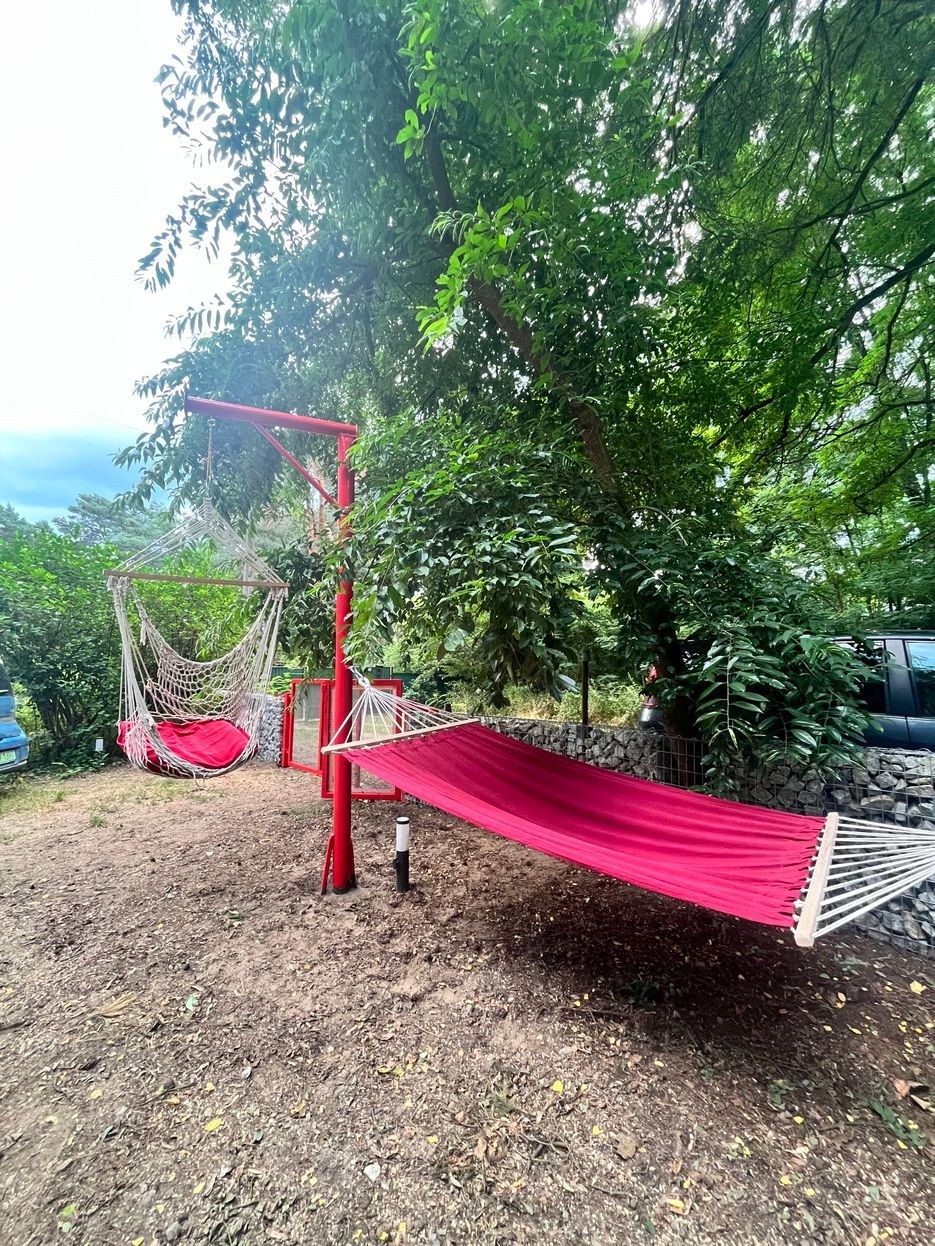 A hammock suspended between red poles in a natural setting with trees and a stone wall.