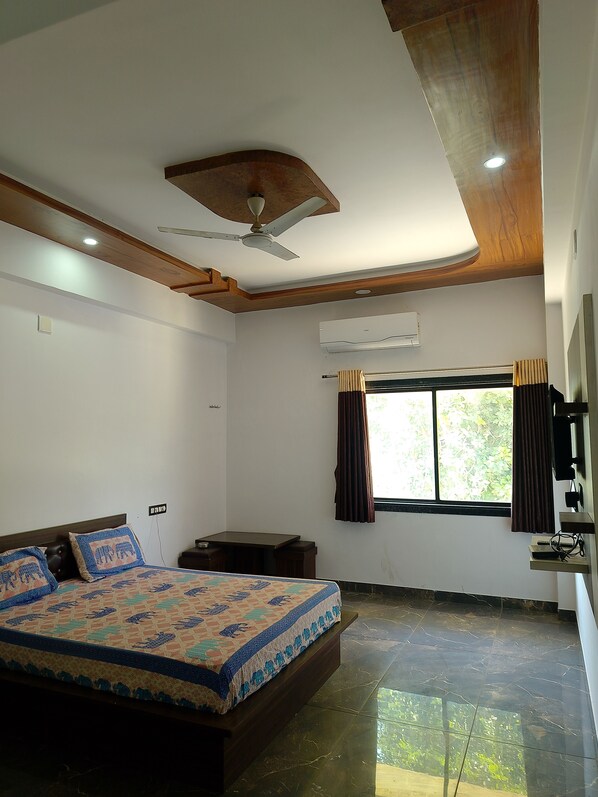 A bedroom with a bed, a ceiling fan, an air conditioning unit, a window with curtains, and a small table. - Hotel Jai Hind (Udaipur)